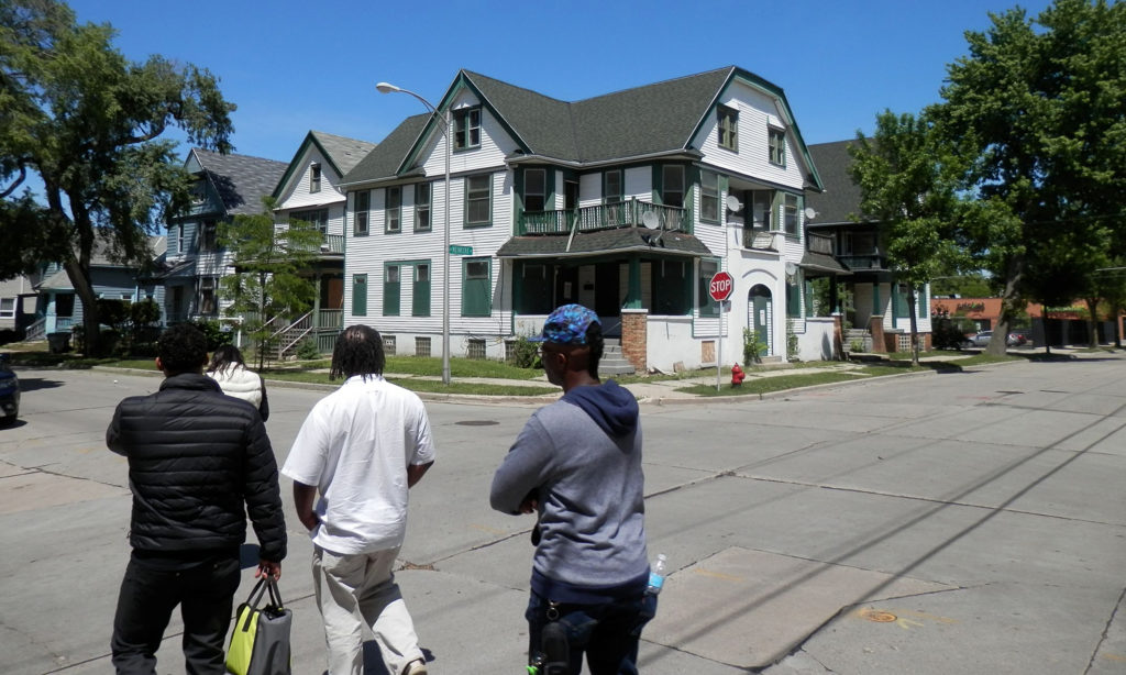 Three people facing away from the camera, looking at the live-work space (a white building with green shutters) in the background.