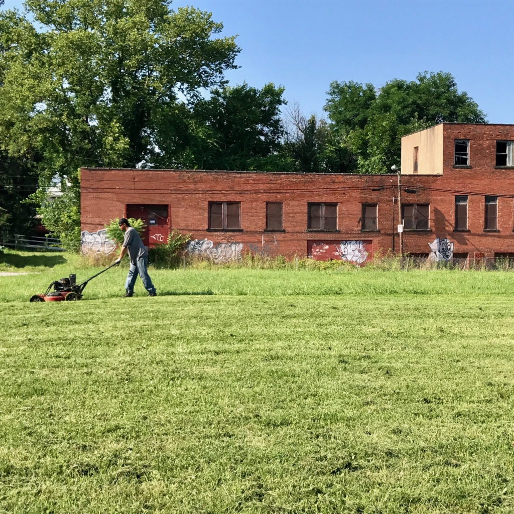 Man mowing a field of grass with an old brick building in the background.