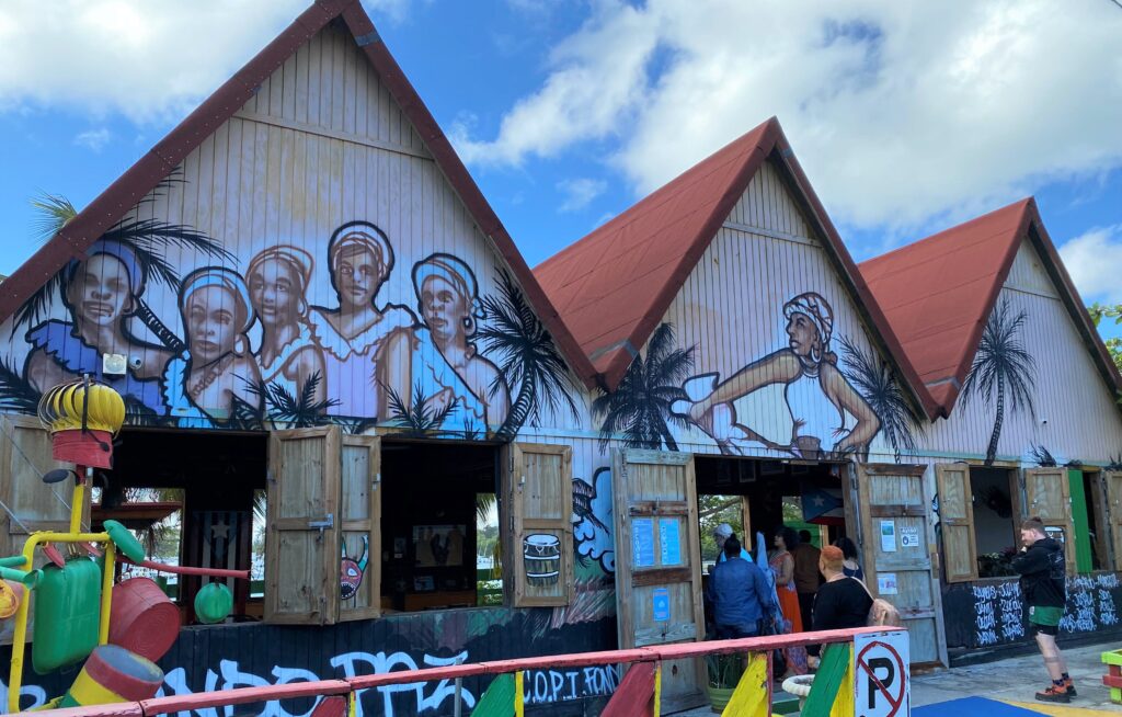 Colorful exterior of Corporación Piñones se Integra (COPI), a community space with three steepled roofs. A mural on the wall of the building depicts Afro-Puerto Ricans and palm trees.
