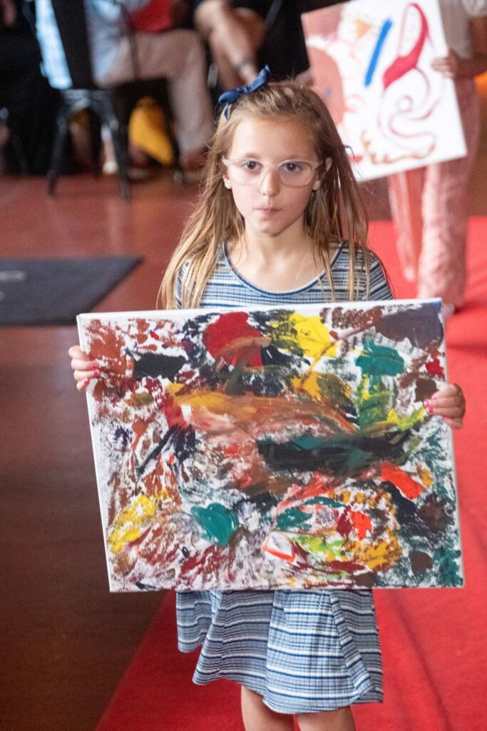 A young white girl walking the runway holding a colorful painting.