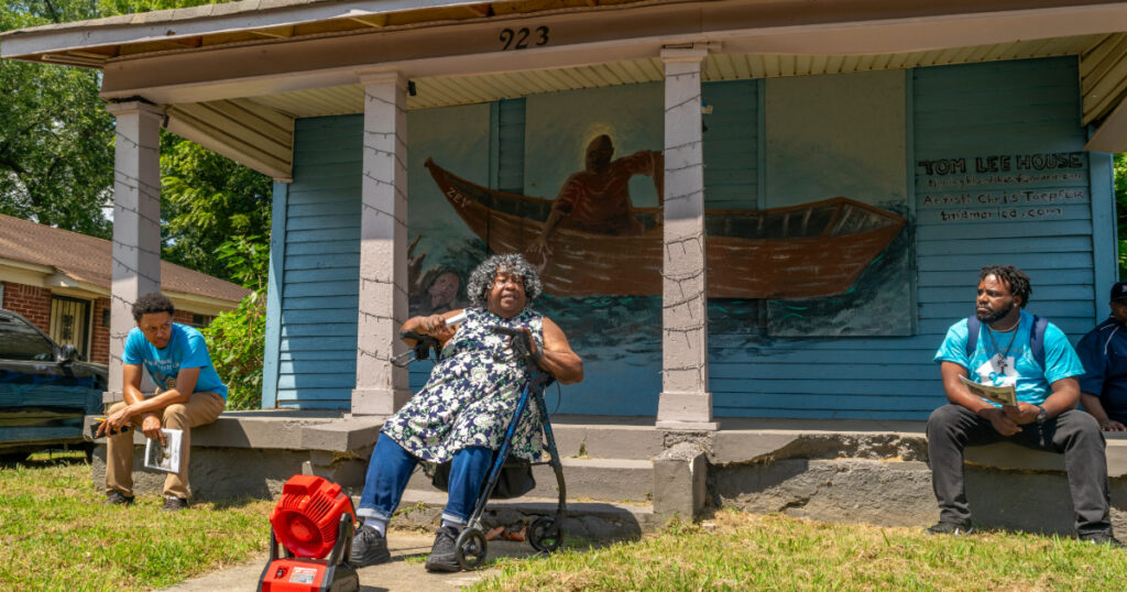 An older Black woman sits in front of the home that once belonged to Tom Lee, a Black riverworker who became a local hero. The home is painted with a mural depicting how he saved the lives of the passengers of a sunken steamboat.