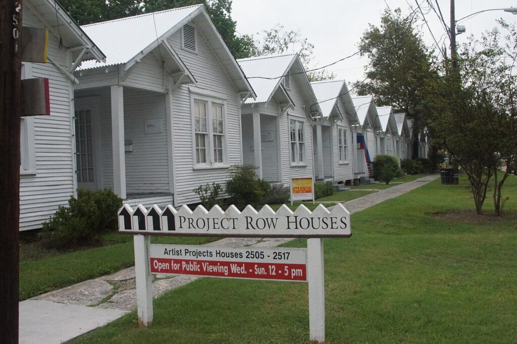 Sign reading "Project Row Houses" in front of a row of several white row houses. 