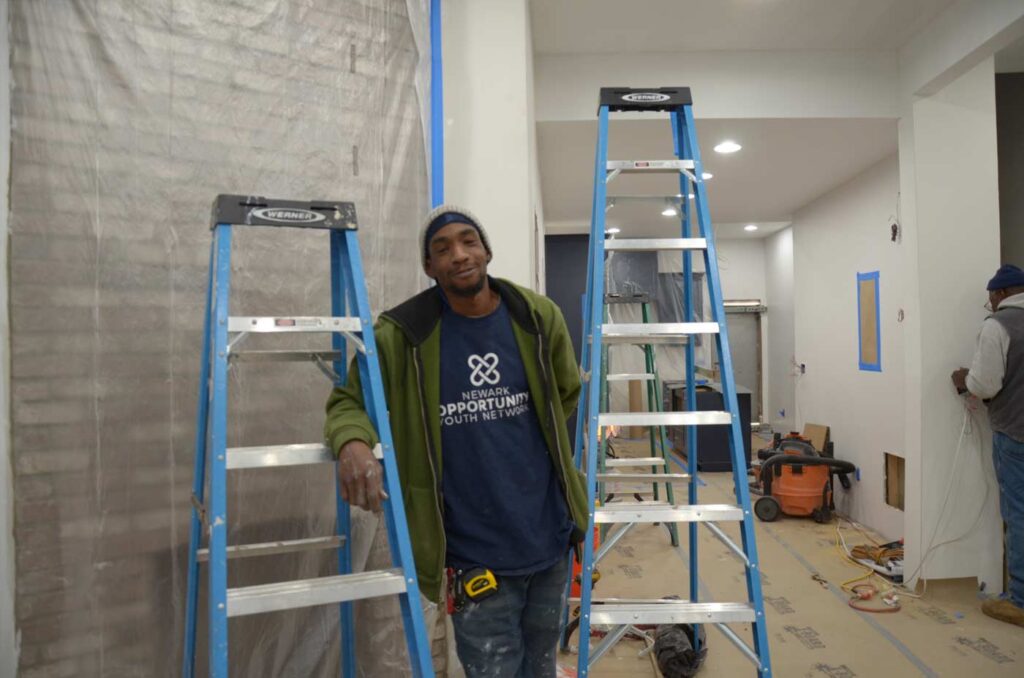 A young Black man in a Youth Build Network t-shirt stans between two ladders.