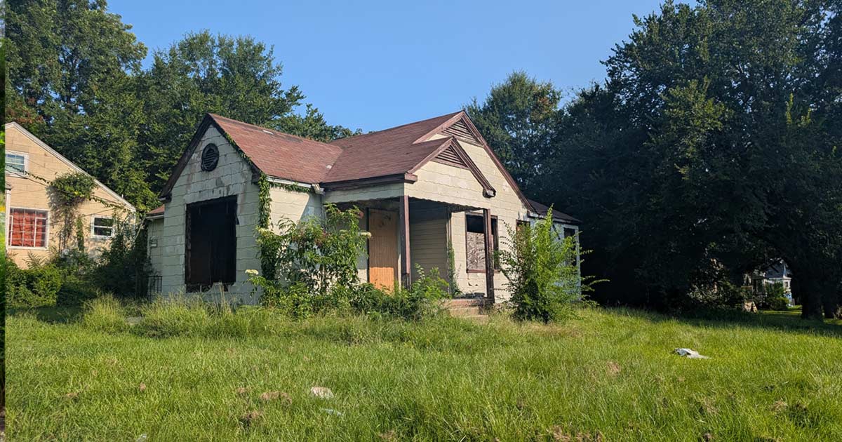 A small vacant bungalow with boarded windows, overgrown vines, and an unmowed lot, surrounded by mature trees on a residential street.