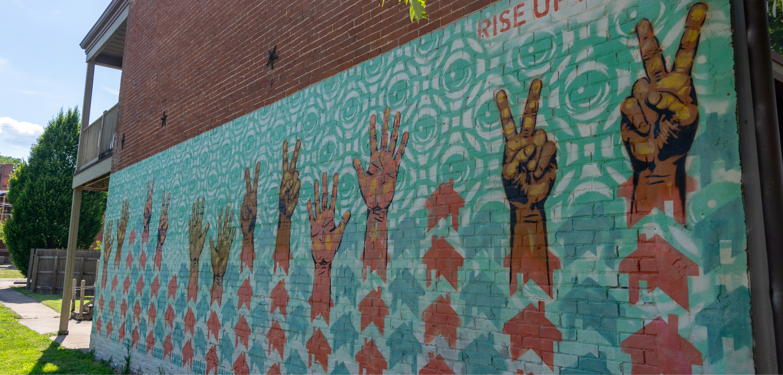 A mural on the side of a building depicting a pattern of blue and red houses, with dark-skinned hands rising from them holding up peace signs and raised fingers. Credit: Center for Community Progress