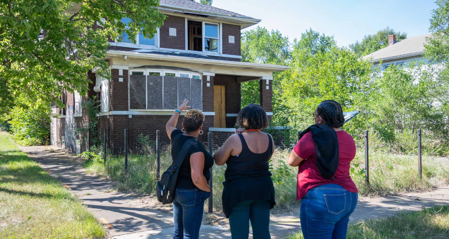 Three women stand in front of a boarded-up single family home, examining it critically
