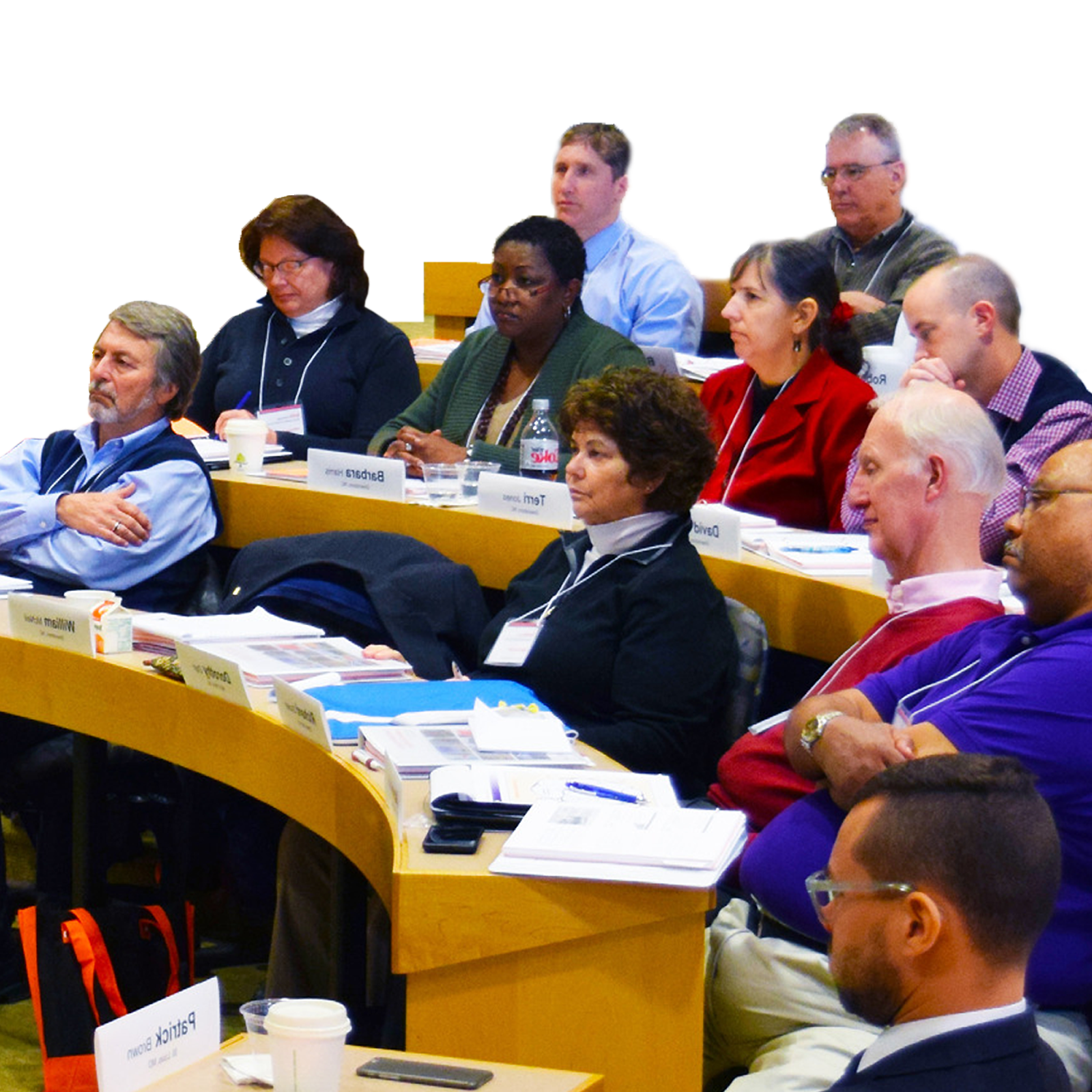A diverse group of attendees seated at curved tables during a conference or workshop, with name placards, notebooks, and materials in front of them.
