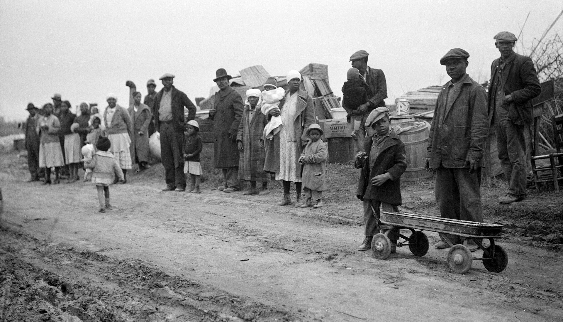 A black-and-white photograph taken in January 1936 by John Vachon showing a large group of Black sharecropper families, including men, women, and young children, standing along a muddy rural roadside in Arkansas after being evicted from their land. Their household belongings — barrels, wooden crates, boxes, and furniture — are piled behind them. A young boy in the foreground holds the handle of a small wagon. The families were displaced by their landowner in retaliation for joining the Southern Tenant Farmers Union.