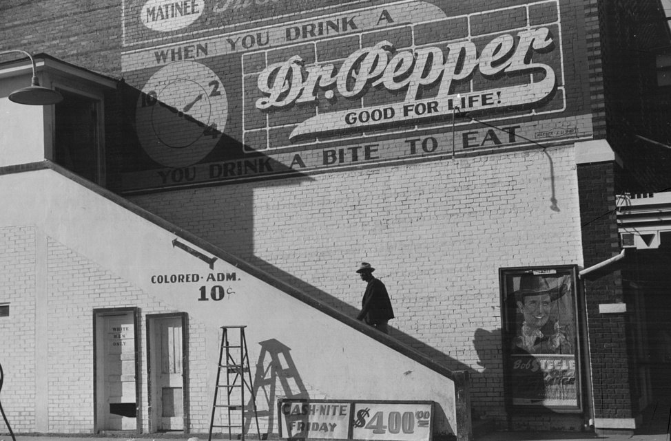 A black-and-white photograph taken in 1939 by Marion Post Wolcott showing the exterior of a movie theater in Belzoni, Mississippi. A Black man climbs an outdoor staircase leading to the segregated upper entrance, marked with a sign reading "Colored Adm. 10¢." At ground level, a door marked "White Men Only" is visible. A large painted Dr. Pepper advertisement covers the upper brick wall, and a movie poster for a Bob Steele film and a "Cash Nite Friday $400" sign are displayed near the entrance. (Source: Marion Post Wolcott / U.S. Library of Congress / Wikimedia Commons)