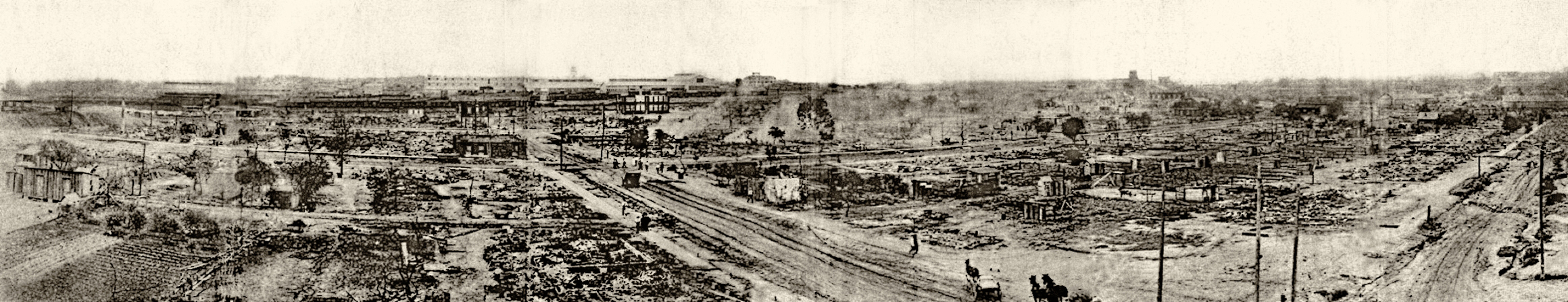 A sepia-toned panoramic photograph showing the vast devastation of the Greenwood District of Tulsa, Oklahoma following the 1921 Tulsa Race Massacre. The image stretches across a wide field of view, revealing block after block of charred rubble, ash, and debris where a prosperous Black neighborhood once stood. Greenwood Avenue runs horizontally through the center of the image. Utility poles, a few scattered structures, and bare trees remain standing amid the destruction. In the far background, larger brick buildings — apparently spared from the destruction — are visible on the horizon. A horse-drawn vehicle and a few figures can be seen on the road. (Source: Mary E. Jones Parrish, "Events of the Tulsa Disaster" / Wikimedia Commons)
