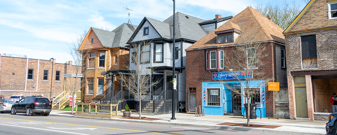 A color photograph of a mixed-use urban block on a Detroit street, showing a row of properties in varying conditions. From left to right: a flat-roofed brick commercial building; a two-story Victorian-style house with warm wood-toned siding under renovation; a tall three-story gray Victorian house with a covered front porch; a brick building with a ground-floor barbershop called "Heavy Weight Cuts" with a bright blue storefront and a barber pole; and a deteriorating brick building at number 4002 with boarded-up windows and doors. Bare trees line the sidewalk and parked cars are visible along the street. The mix of renovated, occupied, and vacant structures illustrates varying stages of disinvestment and reinvestment on a single block.