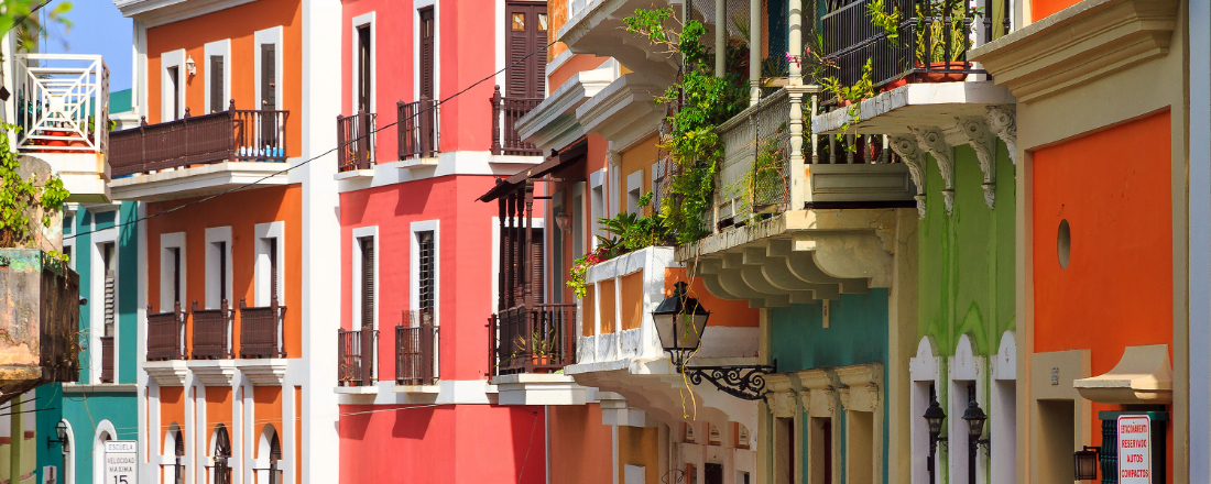 A color photograph of a sunny street in Old San Juan, Puerto Rico, lined with historic multi-story buildings painted in vivid shades of coral pink, orange, teal, and green. The buildings feature ornate white-trimmed balconies with wrought iron railings, wooden shutters, and potted plants spilling over the railings. A decorative cast iron street lamp stands in the foreground, and a speed limit sign is visible at the far left.